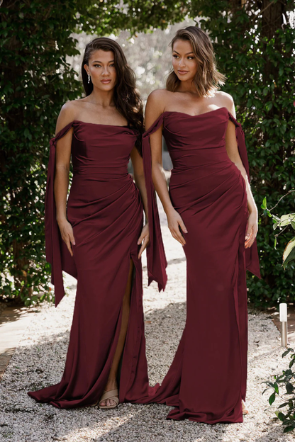 Two women in matching wine strapless slim fit formal, bridesmaid, gala gowns/dresses with a draped design standing outdoors.
