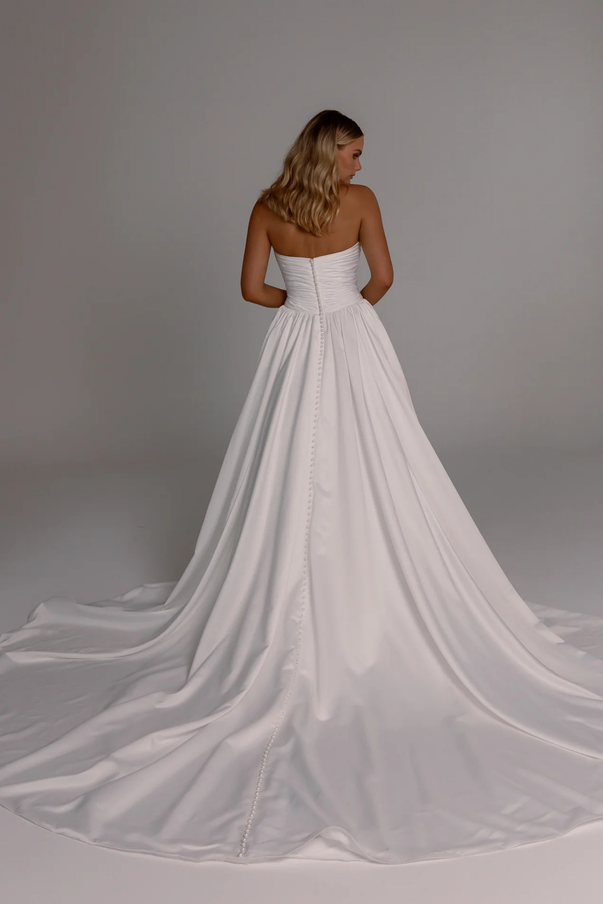 Woman wearing a white strapless wedding dress with a long pearl buttoned train on a plain background