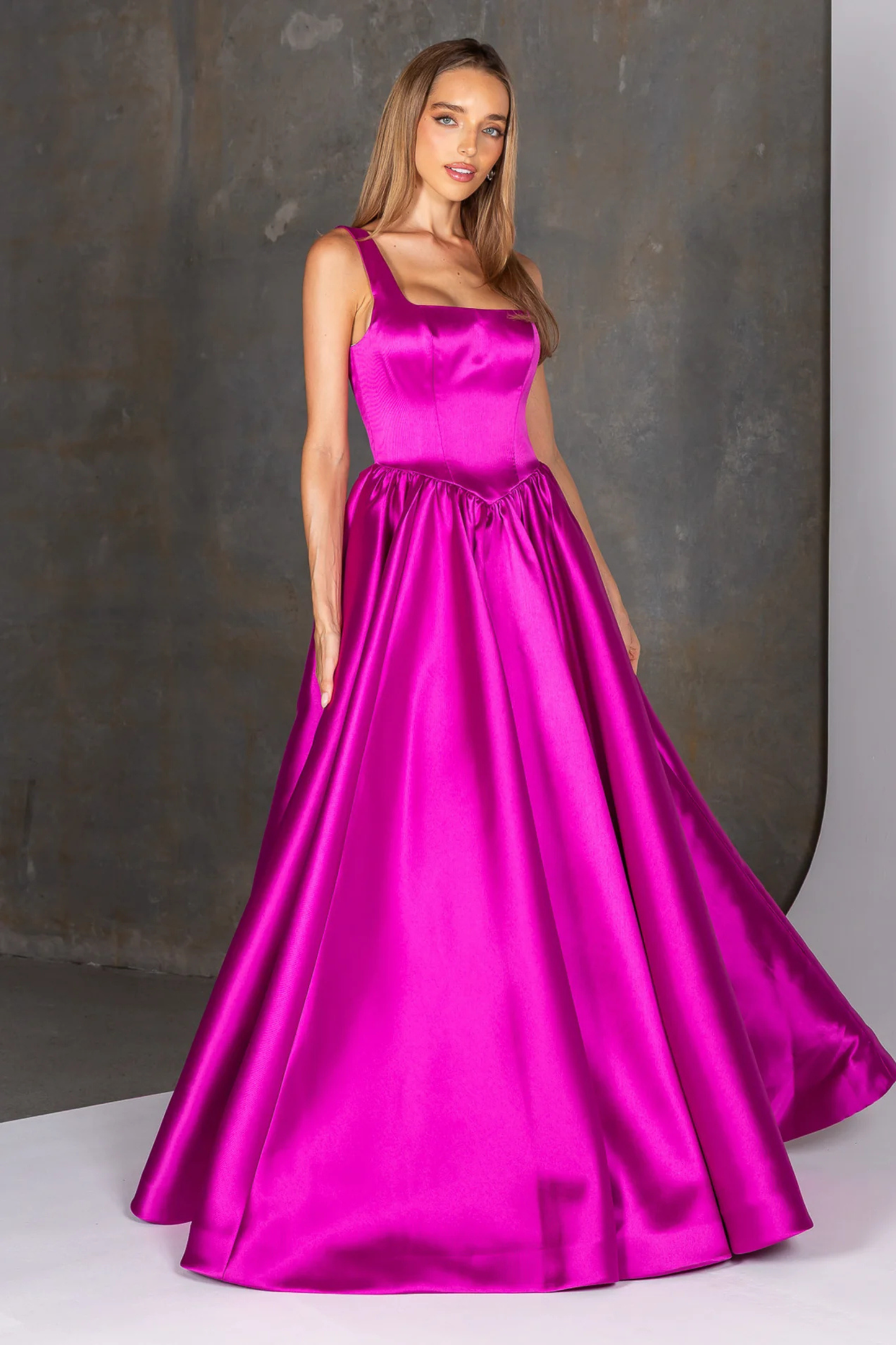 A woman modelling a bright fuchsia Mikado ball gown with a square neckline, wide shoulder straps, a gathered waist, and a full flowing skirt, posed against a dark textured studio backdrop.