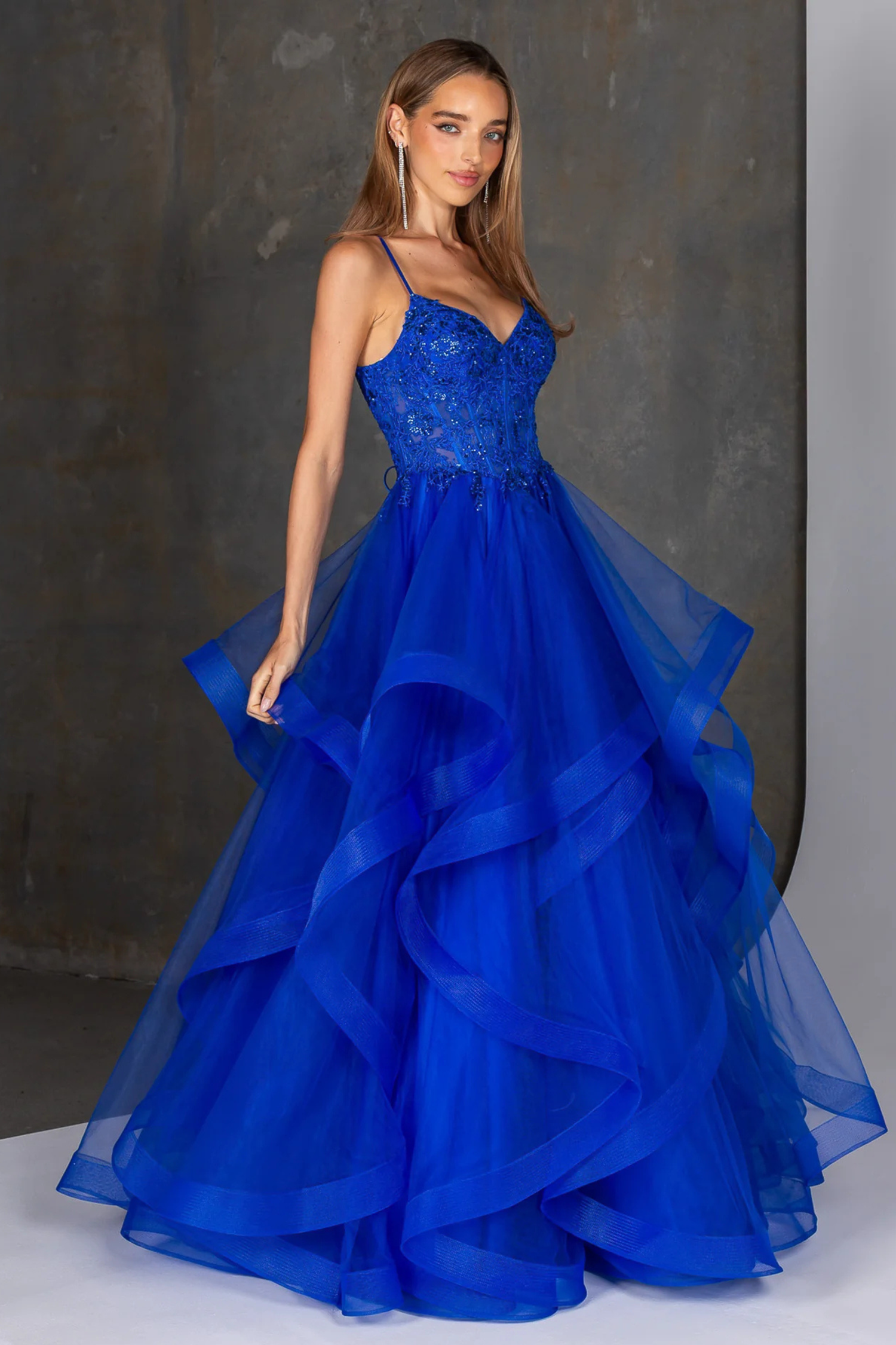 Royal blue evening gown featuring a fitted embellished bodice with delicate beading and thin spaghetti straps, flowing into a dramatic multi-layered tulle skirt with soft cascading ruffles. A statement formal dress ideal for prom, gala, or special occasion wear, showcasing elegant movement and a bold, vibrant color.