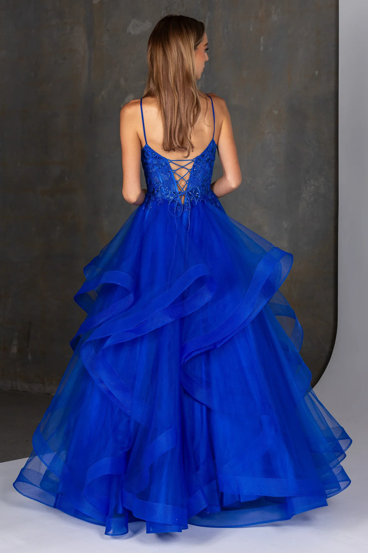 Royal blue evening gown featuring a fitted embellished bodice with delicate beading and thin spaghetti straps, flowing into a dramatic multi-layered tulle skirt with soft cascading ruffles. A statement formal dress ideal for prom, gala, or special occasion wear, showcasing elegant movement and a bold, vibrant color. Showing the back design of the gown.