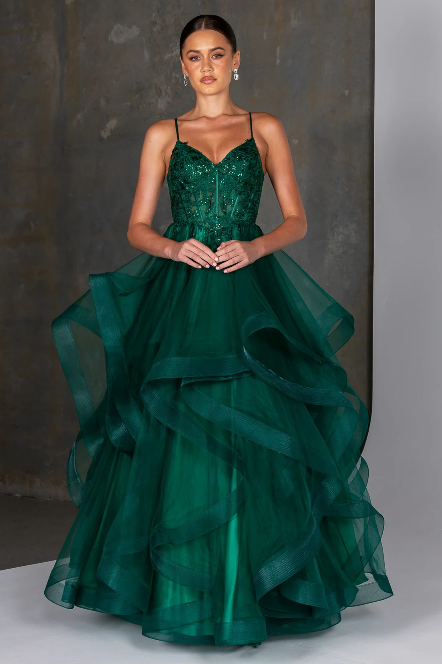 Emerald green evening gown featuring a fitted embellished bodice with delicate beading and thin spaghetti straps, flowing into a dramatic multi-layered tulle skirt with soft cascading ruffles. A statement formal dress ideal for prom, gala, or special occasion wear, showcasing elegant movement and a bold, vibrant color. 