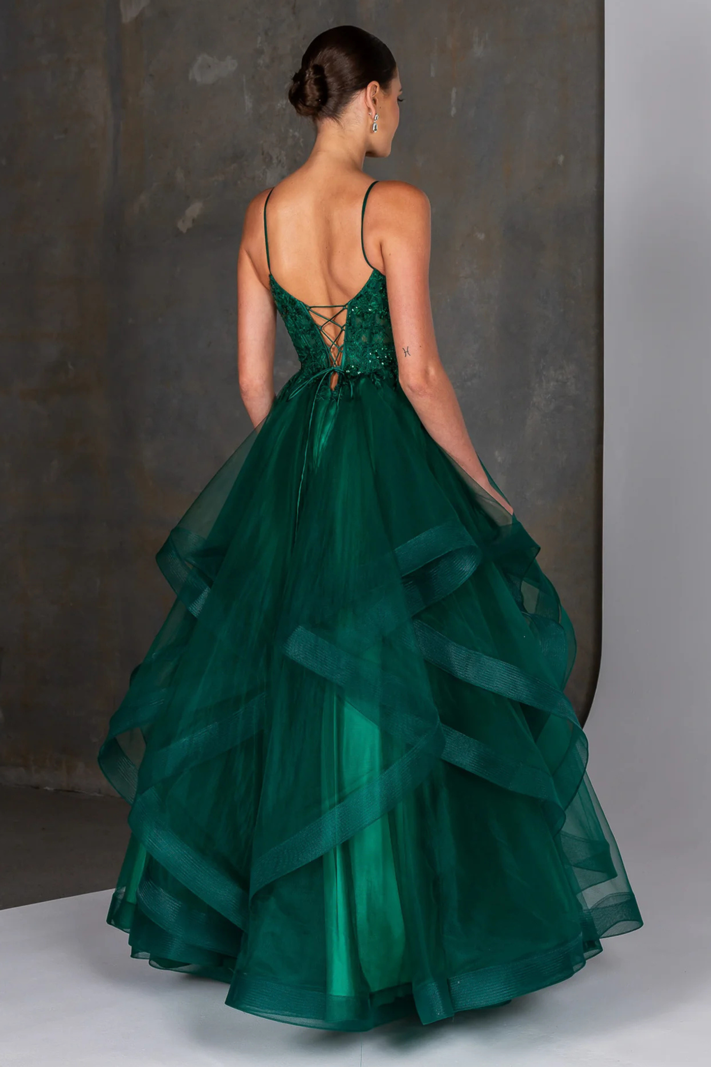 Emerald green evening gown featuring a fitted embellished bodice with delicate beading and thin spaghetti straps, flowing into a dramatic multi-layered tulle skirt with soft cascading ruffles. A statement formal dress ideal for prom, gala, or special occasion wear, showcasing elegant movement and a bold, vibrant color. Showing the back design of the gown.