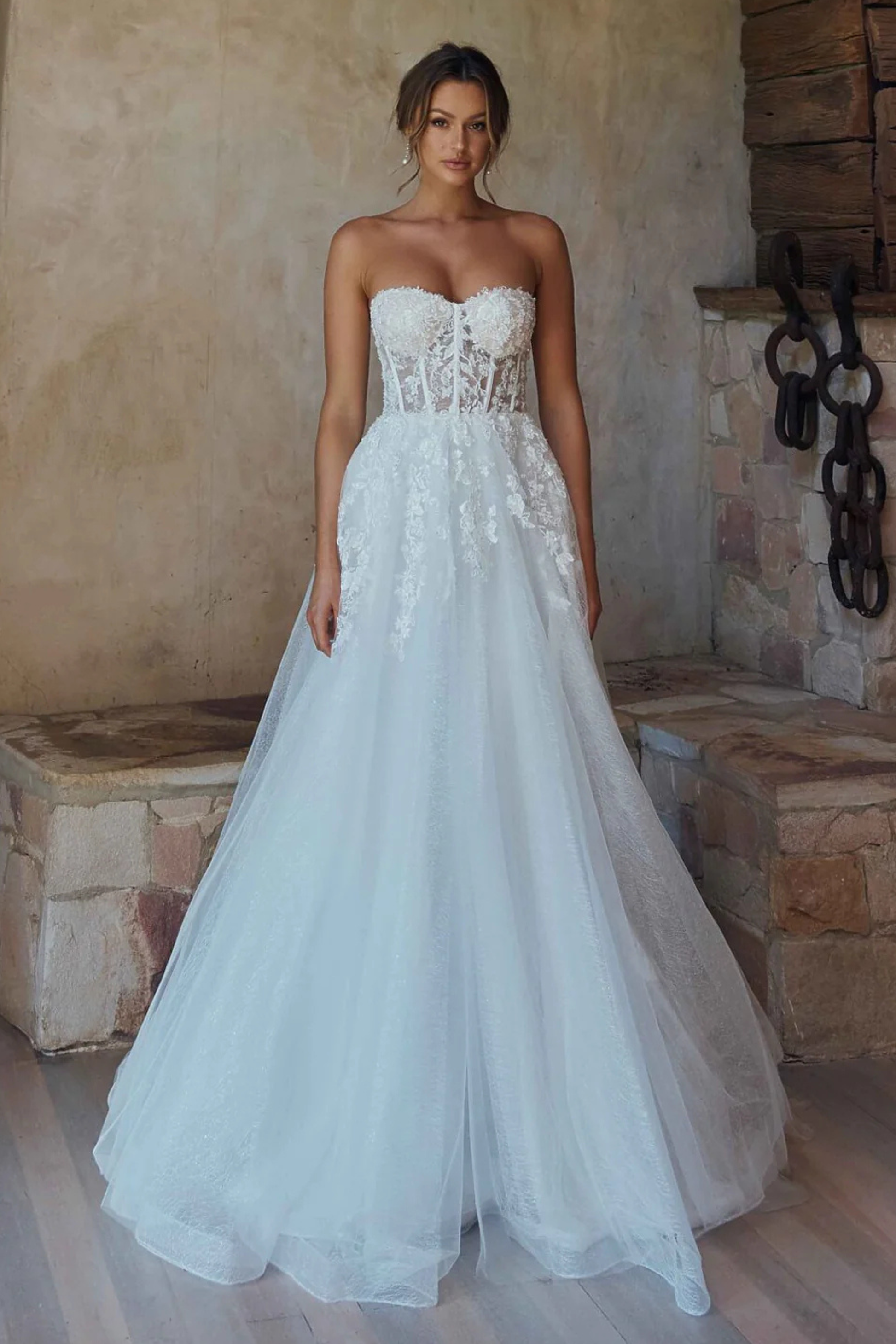 A woman modeling a strapless white wedding gown with a lace-embellished bodice and a full tulle skirt, standing in a rustic stone interior.