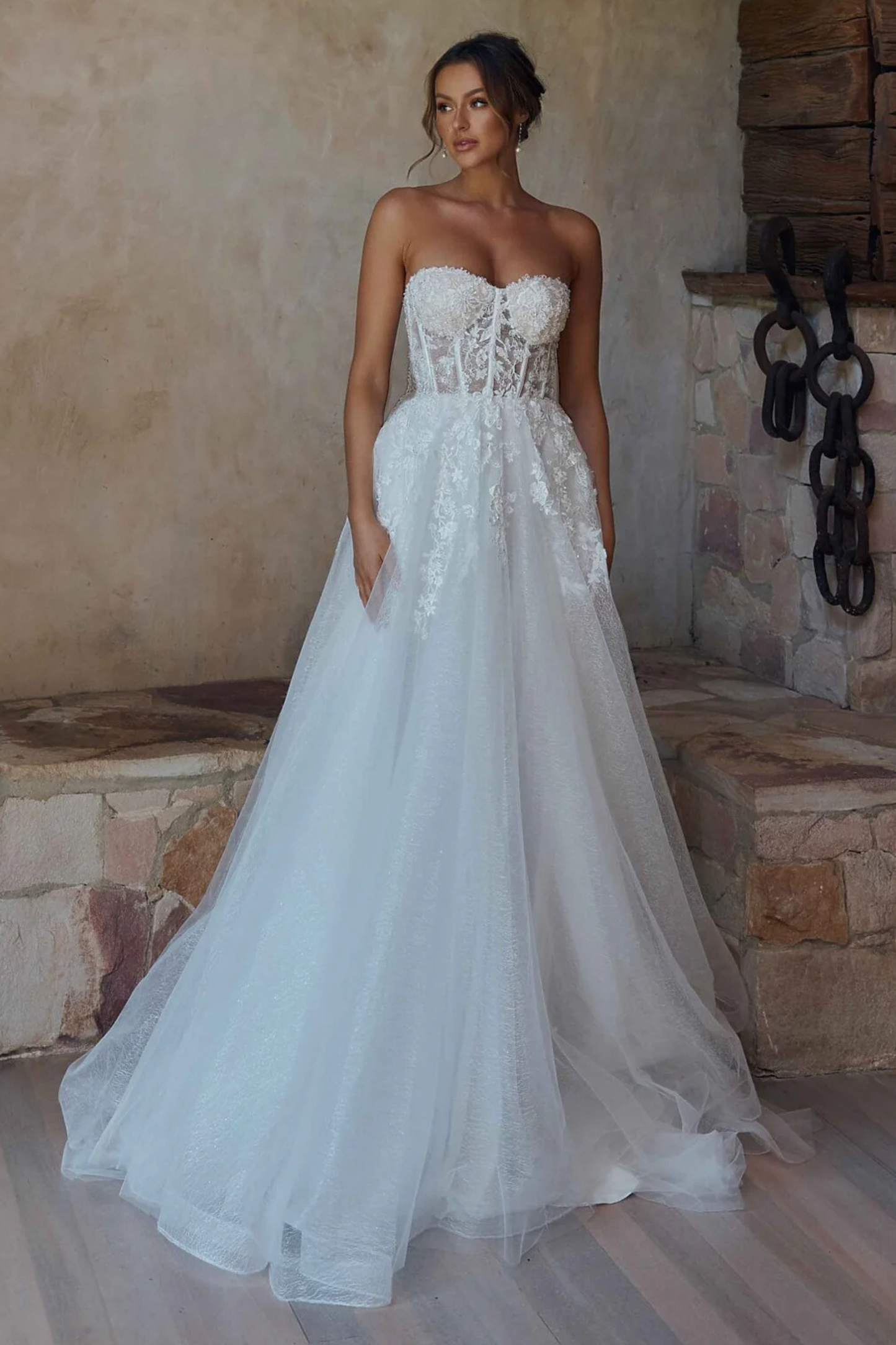 A woman wearing a strapless white wedding gown with a lace-embellished bodice and a full tulle skirt, standing in a rustic stone interior.