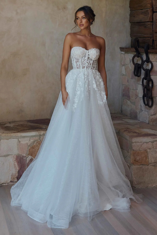 A woman wearing a strapless white wedding gown with a lace-embellished bodice and a full tulle skirt, standing in a rustic stone interior.