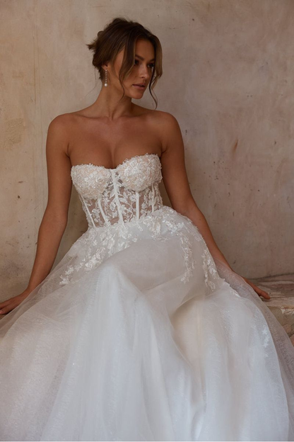 A woman wearing a strapless white wedding gown with a lace corset bodice and full tulle skirt, seated against a textured beige wall.