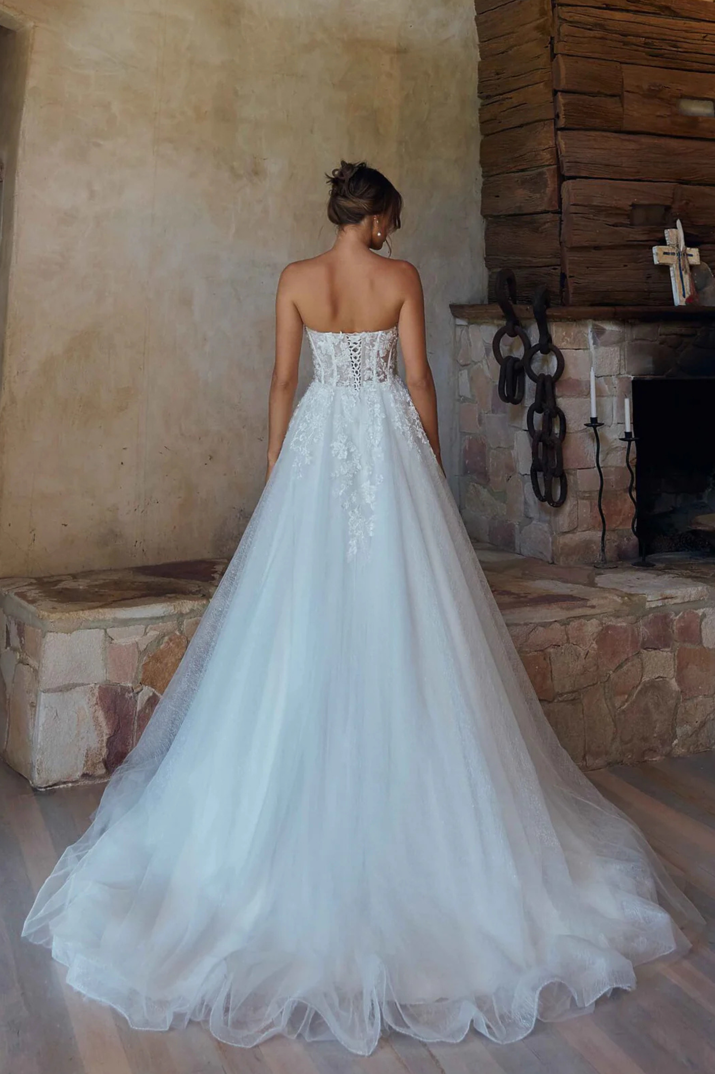 A woman wearing a strapless white wedding gown with lace detailing and a full tulle skirt, photographed from the back in a rustic indoor setting with a stone fireplace and wooden wall.
