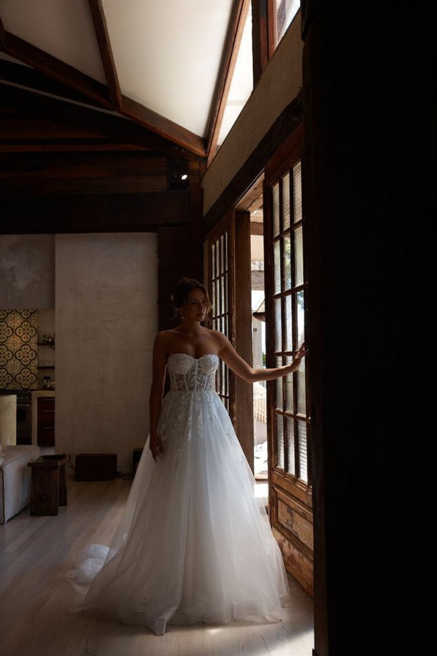 A woman wearing a strapless white wedding gown with a fitted lace bodice and full tulle skirt stands in a dimly lit room near large wooden windows, holding the door frame.