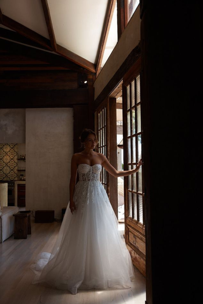 A woman wearing a strapless white wedding gown with a fitted lace bodice and full tulle skirt stands in a dimly lit room near large wooden windows, holding the door frame.