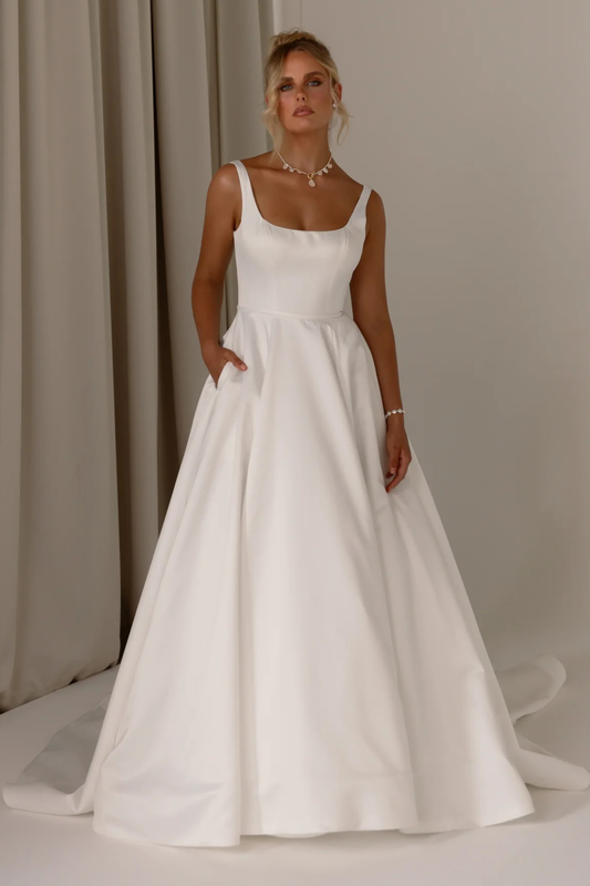 A woman modeling a white A-line wedding dress with thin straps, a square neckline, and side pockets, standing in front of neutral draped curtains.
