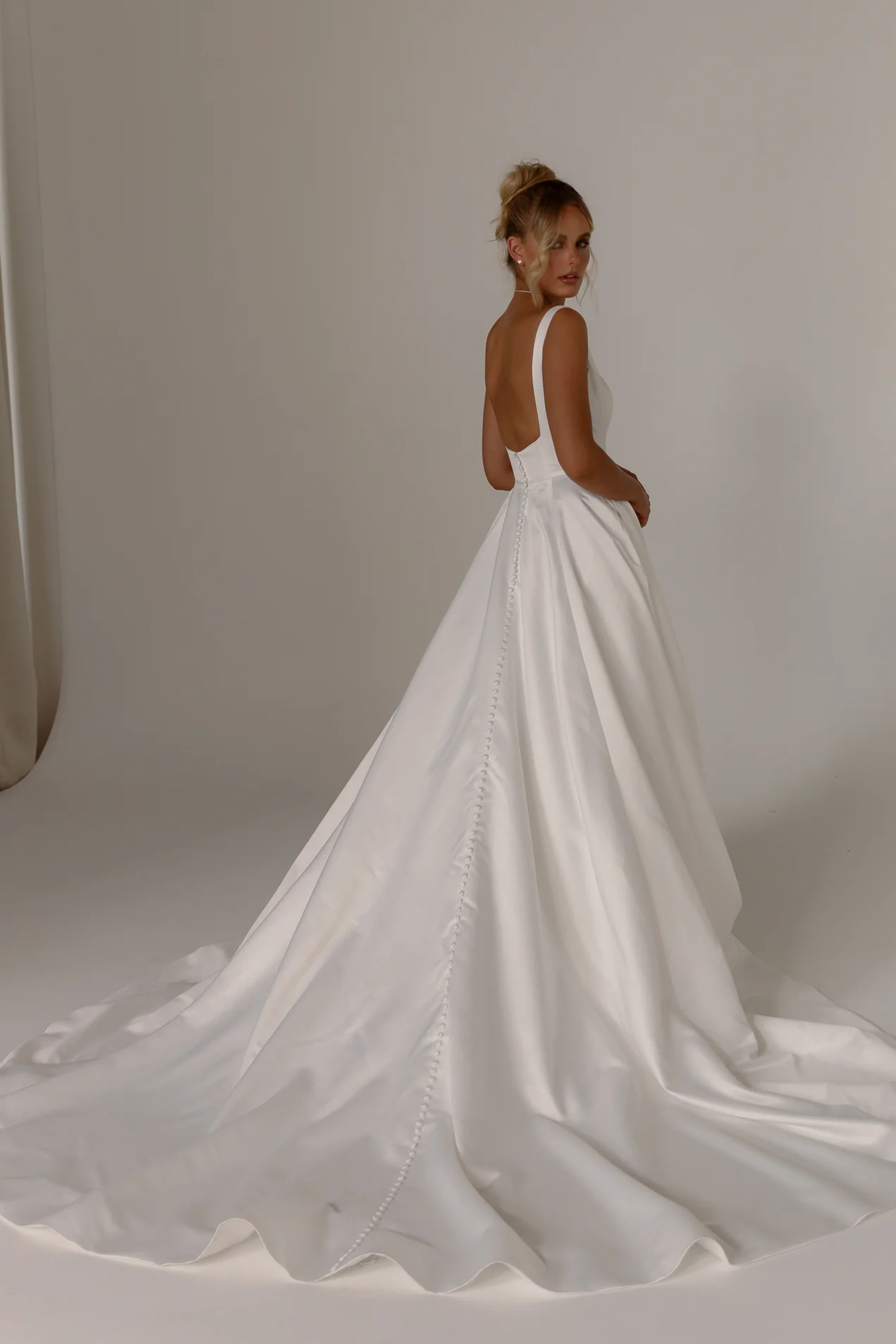A woman modeling a white wedding dress from the back, showing a structured satin-like skirt with a long train, sleeveless straps, and a low open back, photographed in a soft neutral studio setting.